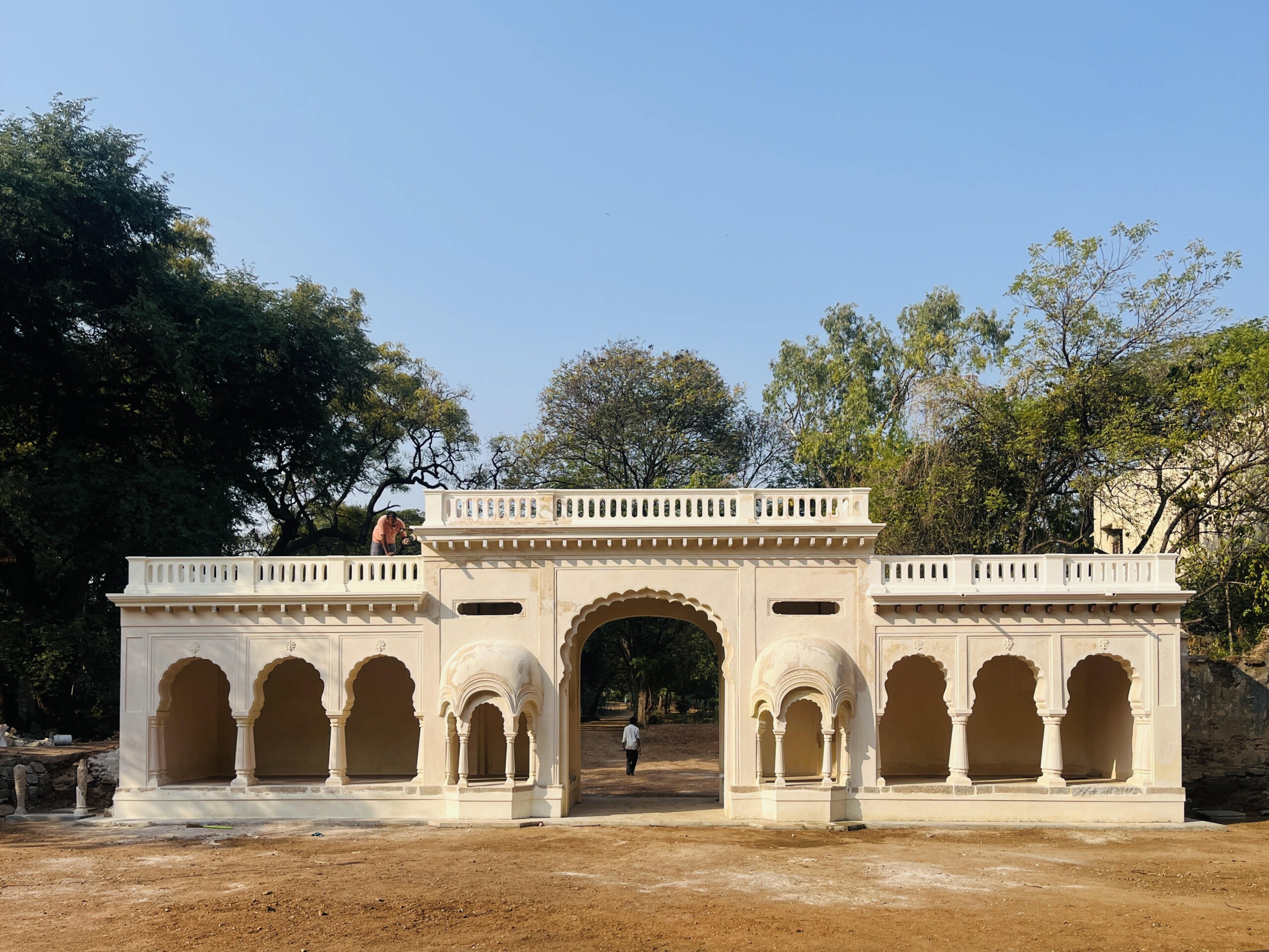 Cream-colored architectural facade with arched doorways and a colonnade, set against a blue sky and surrounding trees; a person stands near the entrance.