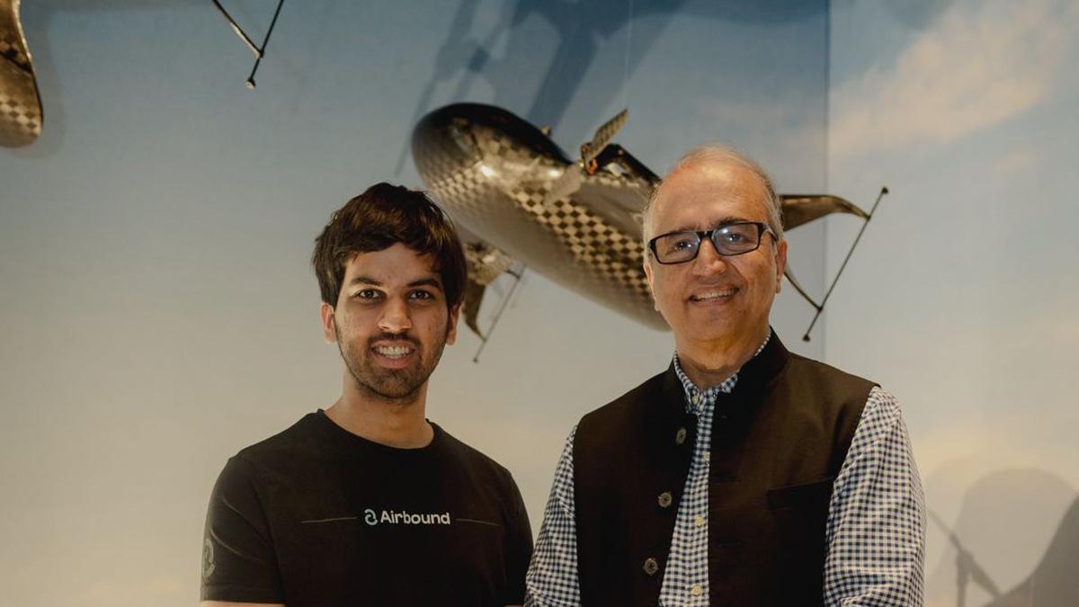 Two men of healthcare industry posing indoors: a younger man in a black Airbound T-shirt and an older man in a checkered shirt with a vest, with a decorative model airplane hanging in the background.