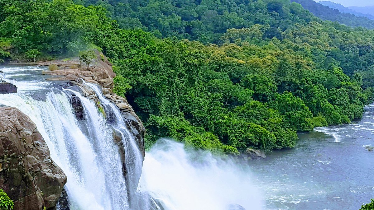Stunning view of Athirappilly Waterfalls, known as Alleppey in Kerala surrounded by dense green forests, flowing river streams, rocky cliffs, and mist rising from the cascading waterfall.