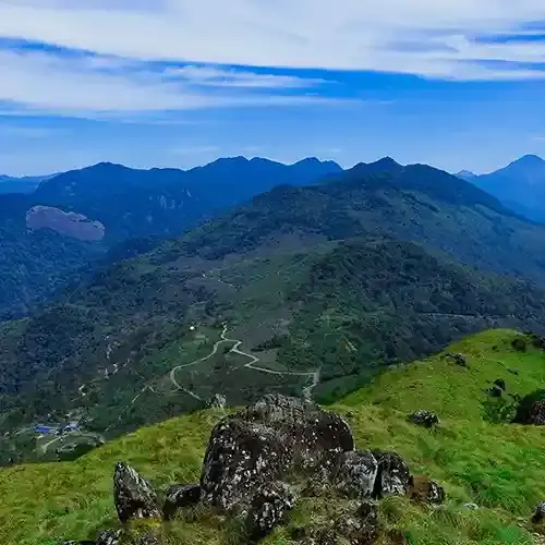 Panoramic mountain landscape of Ponmudi, Kerala, featuring rolling green hills, winding roads, rocky viewpoints, and misty blue mountain ranges under a bright sky.